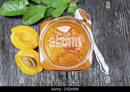 Marmellata di albicocca in vaso di vetro, frutta, un cucchiaio e un ramo con foglie verdi sullo sfondo di legno da sopra Foto Stock
