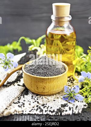 Farina di caraway nero in una ciotola, semi in un cucchiaio di burlap, olio in bottiglia e ramoscelli Nigella sativa con fiori blu e foglie su legno bordo backgro Foto Stock