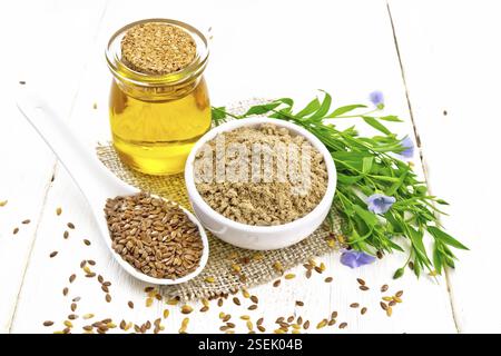 Farina di semi di lino in una ciotola, semi di lino in un cucchiaio, olio in vaso di vetro su un tovagliolo di tela, foglie e fiori di lino blu su sfondo di legno chiaro Foto Stock
