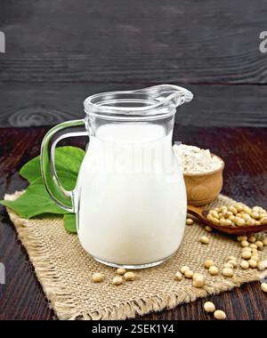 Soya il latte in una caraffa, infarinare in una ciotola e foglia verde, soia in un cucchiaio e baffè sullo sfondo di una tavola di legno scuro Foto Stock