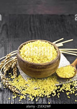 Bulgaro groats - grani di grano al vapore - in una ciotola di argilla e un cucchiaio sul insaccamento, orecchie di grano sullo sfondo di tavola di legno Foto Stock