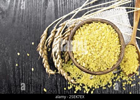Bulgaro groats - grani di grano al vapore - in una ciotola di argilla e cucchiaio su un tovagliolo di burlap, orecchie di grano su sfondo di legno da sopra Foto Stock