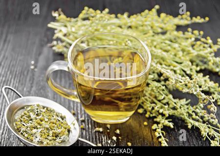 Tisana di wormwood grigio in una tazza di vetro, fiori freschi e setaccio metallico con fiori secchi paginetta su sfondo di legno Foto Stock