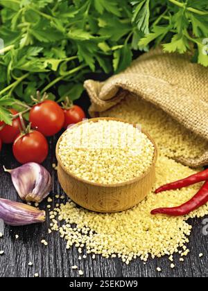 Couscous crudo in una ciotola e un sacco di burlap, aglio, pomodori, peperoni caldi e erbe sullo sfondo di legno scuro tavola Foto Stock