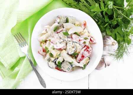 Insalata con zucchine fritte, uova sode, pomodoro fresco e aglio, condito con maionese o panna acida in un piatto, tovagliolo e prezzemolo su tavola di legno Foto Stock