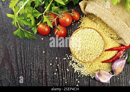 Couscous crudo in una ciotola e un sacco di sacco, aglio, pomodori, peperoncini ed erbe aromatiche su uno sfondo di legno nero dall'alto Foto Stock