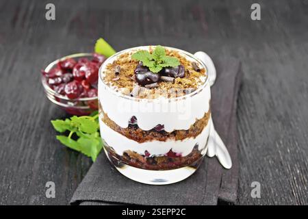 Dessert Foresta Nera di ciliegie, biscotto al cioccolato e formaggio casolare morbido con panna in un bicchiere su tovagliolo, menta, bacche in una ciotola e un cucchiaio su bl Foto Stock