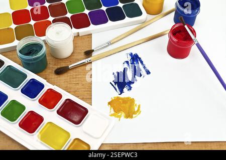 Due scatole di vernice, tre spazzole, vasi con gouache, un foglio di carta con un'immagine su una tavola di legno Foto Stock