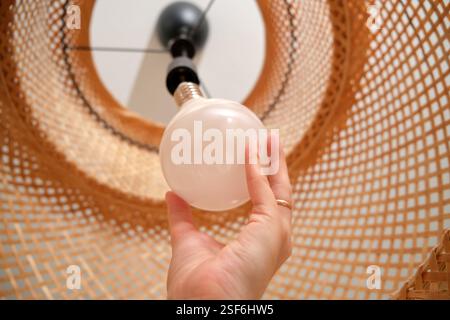 Tenere la lampadina bianca mentre la si installa in un paralume in rattan intrecciato. Primo piano della configurazione dell'illuminazione domestica. Foto Stock