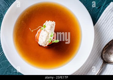 Zuppa o consomme, brodo di carne denso con carote, gnocchi di pollo e microverdure, servito in un piatto bianco su sfondo verde scuro con un tovagliolo A. Foto Stock