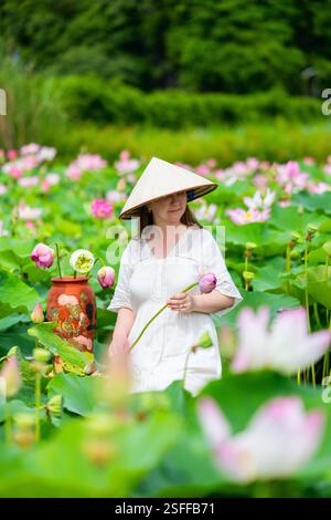 Una donna che indossa un tradizionale cappello conico vietnamita regge un fiore di loto rosa mentre è in piedi in un vivace campo di fiori di loto e foglie verdi Foto Stock