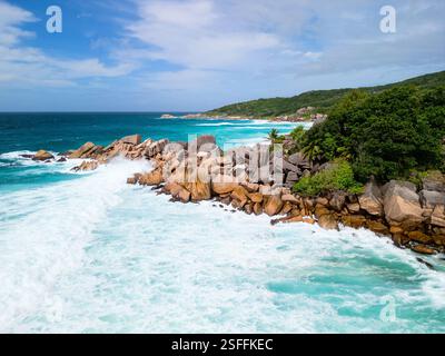 Una vista aerea panoramica di la Digue, Seychelles, che mostra onde turchesi che si infrangono contro formazioni rocciose di granito uniche e lussureggiante vegetazione tropicale Foto Stock
