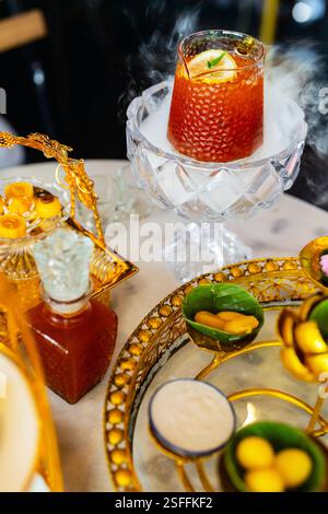 Un ambiente decorativo caratterizzato da un bicchiere di tè al limone ghiacciato su ghiaccio secco, che crea un effetto fumé, circondato da una varietà di dolci tradizionali e ornati Foto Stock