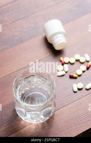 Un bicchiere d'acqua accanto alle pillole versate su una superficie di legno. Il flacone è rovesciato, suggerendo un uso recente. La scena trasmette temi di salute e m Foto Stock