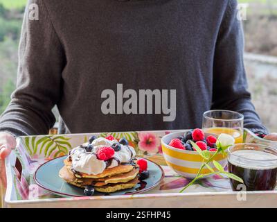 Uomo che porta una colazione di San Valentino su un vassoio Fiori, espresso, pancake e frutta rossa Foto Stock