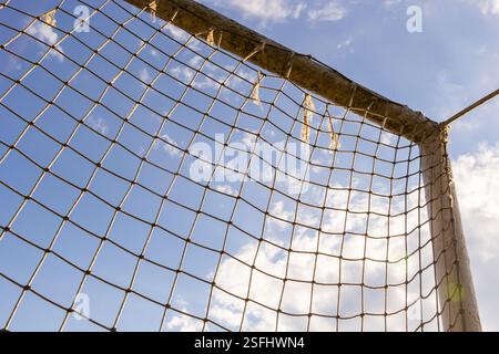 Un'antica rete da calcio si erge alta con fili sfilacciati, contro un cielo blu vibrante punteggiato da nuvole morbide, a simboleggiare il gioco all'aperto e lo sport Foto Stock
