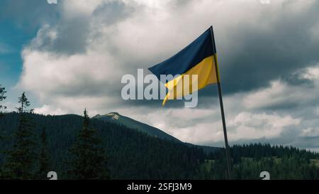 The Ukrainian flag waves proudly against the backdrop of forested mountains and dramatic skies. Foto Stock
