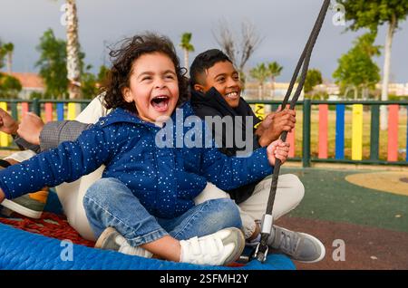 Fratello e sorella ridono ad alta voce mentre sfrecciano su un parco giochi, trascorrendo una bella giornata all'aperto con la famiglia Foto Stock