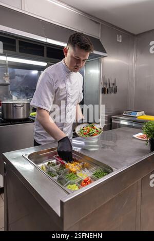 Lo chef utilizza pinze e tiene il piatto con insalata fresca sopra i contenitori con ingredienti nella cucina del ristorante Foto Stock