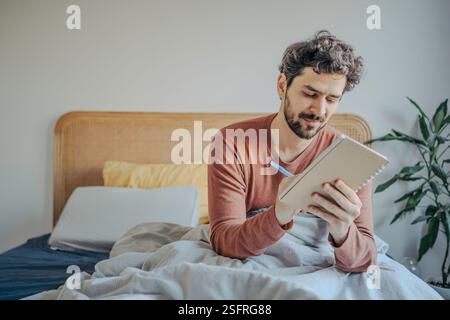 Giovane bell'uomo medio-orientale seduto a letto e a scrivere sul suo quaderno. Creatività mattutina e riflessione. Foto Stock
