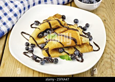Due pancake con mirtilli, menta e glassa di cioccolato su un piatto, tovagliolo, ciotola su uno sfondo di tavole di legno Foto Stock