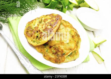 Frittelle di zucchini, aneto e prezzemolo in una piastra con un panno da cucina, panna acida nel piattino sulla tavola di legno sfondo Foto Stock
