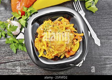 Pollo stufato con pomodori, peperoni gialli e rossi e formaggio in un piatto su tovagliolo, timo, prezzemolo e aglio su fondo di legno nero Foto Stock