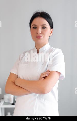 Cosmetologa sicura di sé in camice da laboratorio in piedi con le braccia incrociate in clinica. Il giovane e bellissimo professionista caucasico sta guardando la macchina fotografica al salone Foto Stock