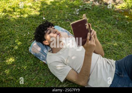 Uno studente sdraiato sull'erba, a leggere un libro nel tranquillo verde del campus. Ideale per temi di educazione, relax, vita studentesca e vita all'aperto Foto Stock