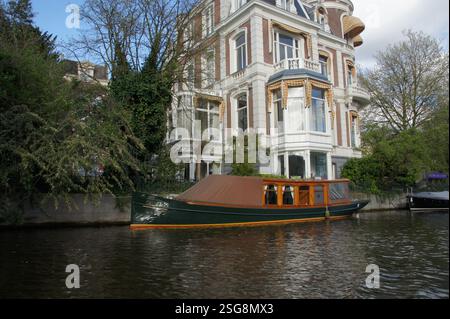 Una casa galleggiante galleggia su un canale panoramico di Amsterdam fiancheggiato da alberi. La tranquilla scena si snoda lungo Singelgracht, un'affascinante zona residenziale. Foto Stock