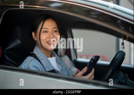 Una donna asiatica positiva che tiene il suo smartphone mentre guida, guarda fuori dalla finestra. tecnologia wireless, trasporti, stile di vita Foto Stock