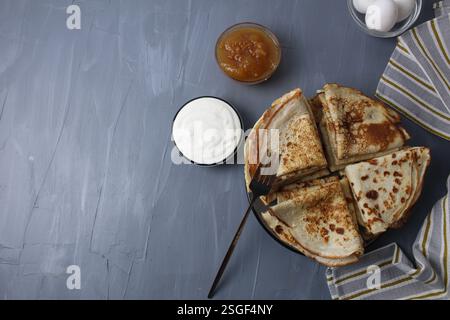 Crepe pancake piegati in triangoli distesi su un piatto con panna acida e marmellata in barca su sfondo grigio con spazio per il testo. Foto Stock