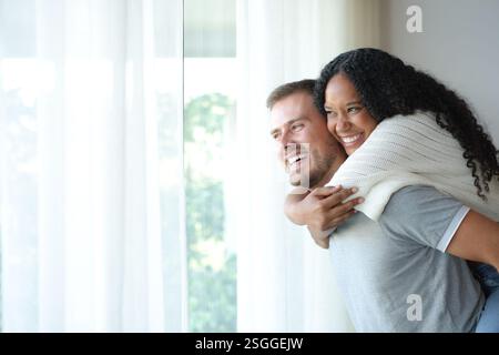 Felice coppia interrazziale che scherza guardando da una finestra a casa Foto Stock