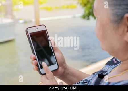 Anziana donna asiatica matura seduta su sedia vicino al fiume e utilizzando il telefono cellulare. Messa a fuoco selettiva, vista spalla. Foto Stock