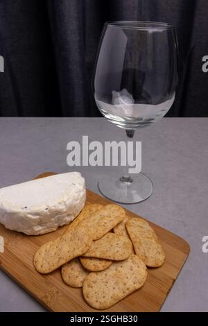 Un vassoio di legno contiene formaggio morbido e cracker croccanti accanto a un bicchiere trasparente. Lo sfondo è scuro ed elegante. Foto Stock