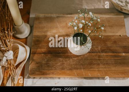 Un vaso di fiori bianchi siede su un tavolo di legno. Il tavolo è ricoperto da un panno bianco e un tovagliolo bianco. Il vaso si trova al centro del ta Foto Stock