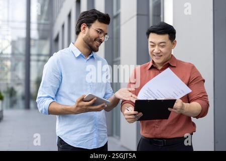 Due colleghi di dipendenti di uffici che discutono relazioni finanziarie, contratti e documenti, un gruppo di uomini d'affari all'esterno dell'edificio con documenti in mano che discutono. Foto Stock