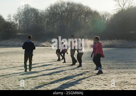 Un gruppo di sei persone fa jogging su un campo coperto di gelo in una mattina fredda, con il sole che getta lunghe ombre e crea una nebbia d'atmosfera. Sale Water Park, Manchester, Regno Unito Foto Stock