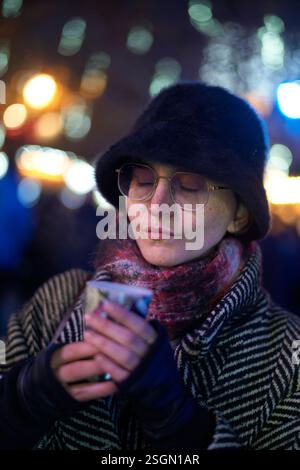 Donna con un'intima sciarpa e cappello che sorseggia una bevanda calda in un mercato notturno di festa. Foto Stock