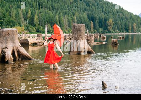Donna con un vestito rosso e un cappello che tiene in mano un ombrello in piedi in un panoramico lago nella foresta. Rattlesnake Lake, Puget Sound, Washington, USA Foto Stock