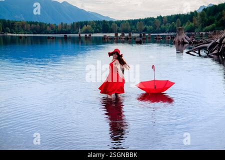 Donna in abito rosso e cappello si trova nel lago vicino a un ombrello rosso, circondato da montagne e alberi. Rattlesnake Lake, Puget Sound, Washington, USA Foto Stock
