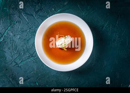 Zuppa o consomme, brodo di carne denso con carote, gnocchi di pollo e microverdure, servito in un piatto bianco su sfondo verde scuro con cucchiaio. Vie Foto Stock