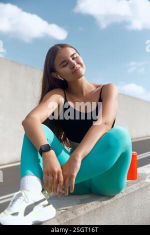 Una giovane donna si siede su una sporgenza in cemento indossando abbigliamento sportivo, sorridendo e ascoltando musica tramite auricolari wireless, godendosi un momento di relax all'aperto Foto Stock