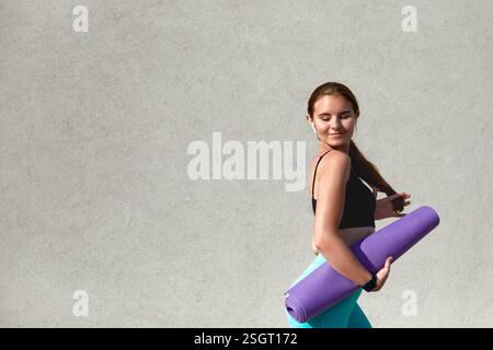 Una giovane donna dai capelli lunghi regge un tappetino yoga viola, indossa una canotta nera e leggings azzurri. Sembra rilassata e sicura di sé mentre si appresta Foto Stock