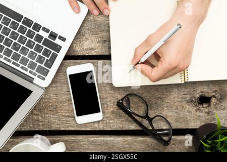 Persona che scrive appunti mentre utilizza un computer portatile e un telefono cellulare su un tavolo in legno rustico durante una sessione di lavoro Foto Stock