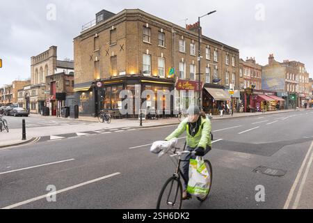 Ciclista su Stoke Newington Church Street, Londra, Regno Unito, vicino a un negozio all'angolo. Una persona guida una bicicletta carica di borse. Foto Stock