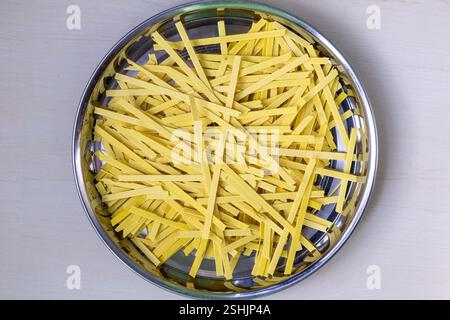 Spaghetti piatti crudi su una piastra di acciaio, sparati dall'alto. Sfondo in legno chiaro. Foto Stock