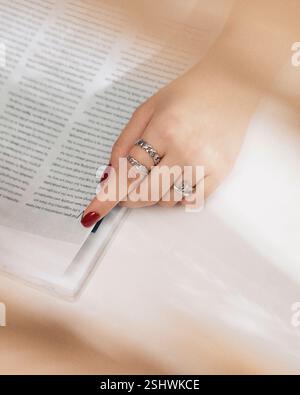 Primo piano della mano di una donna con smalto rosso che gira una pagina in una rivista, mostrando eleganti anelli in argento Foto Stock