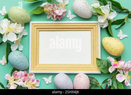 Uova di Pasqua, fiori primaverili, farfalle e una cornice vuota creano un bordo festoso su uno sfondo turchese Foto Stock