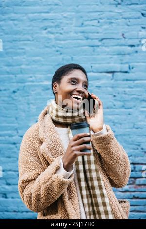 La giovane donna allegra sta parlando al telefono cellulare e tiene in mano una tazza di caffè mentre si trova contro un muro blu Foto Stock